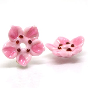 May include: Two pink ceramic flower beads with brown centers. The beads are shaped like five-petal flowers with a hole in the center.