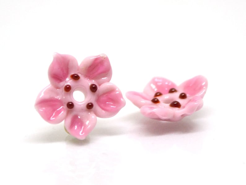 May include: Two pink ceramic flower beads with brown centers. The beads are shaped like five-petal flowers with a hole in the center.