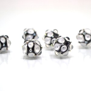 May include: Six white and black glass beads with a geometric pattern. The beads have a shiny, iridescent finish.