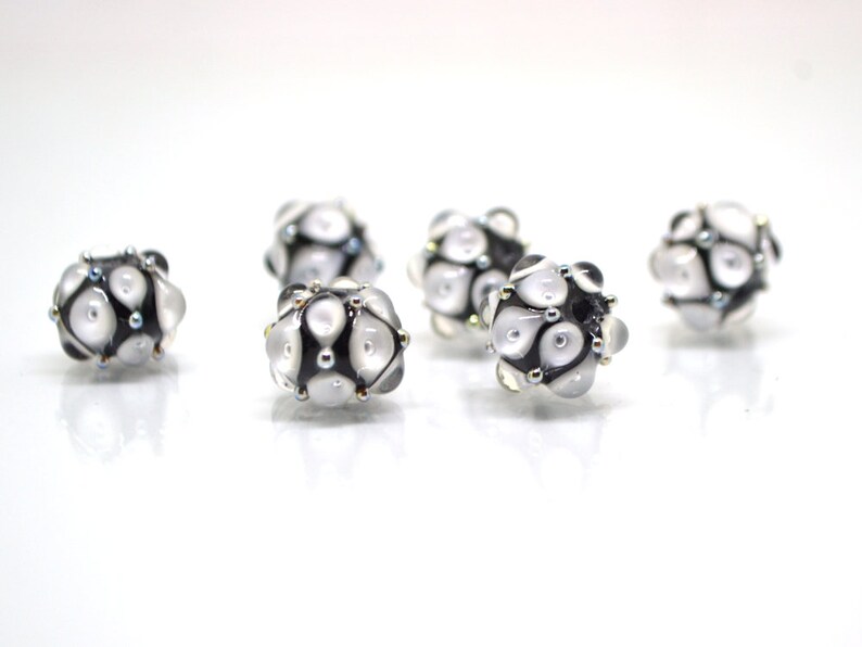 May include: Six white and black glass beads with a geometric pattern. The beads have a shiny, iridescent finish.