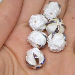 May include: A collection of six white glass beads with iridescent blue and purple accents. The beads are shaped like small flowers with a textured surface.