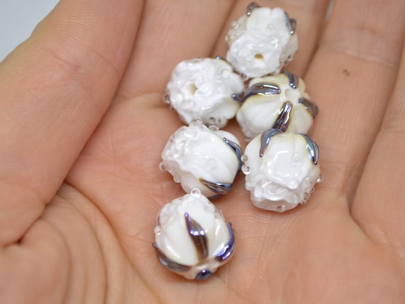 May include: A collection of six white glass beads with iridescent blue and purple accents. The beads are shaped like small flowers with a textured surface.