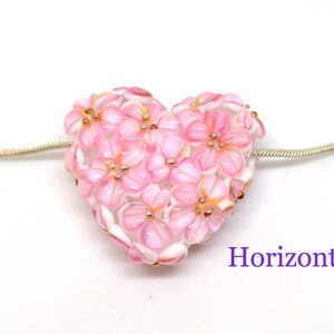 May include: A pink glass heart charm with a floral design. The charm is attached to a silver chain.