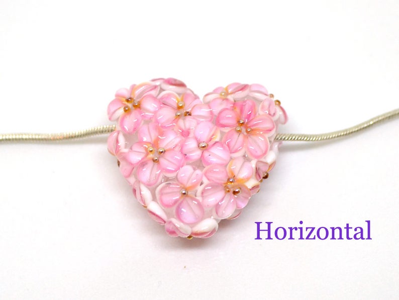 May include: A pink glass heart charm with a floral design. The charm is attached to a silver chain.