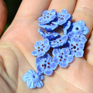 May include: A collection of blue glass flower beads with red centers. The beads are arranged in a pile on a hand.