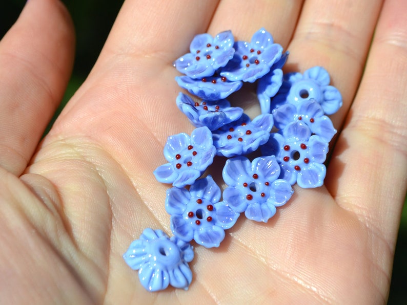 May include: A collection of blue glass flower beads with red centers. The beads are arranged in a pile on a hand.