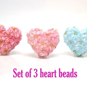 May include: Set of three heart-shaped beads. Two are pink with white flowers and one is light blue with white flowers and gold accents.