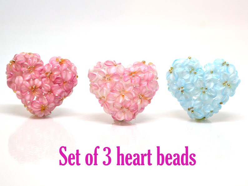 May include: Set of three heart-shaped beads. Two are pink with white flowers and one is light blue with white flowers and gold accents.