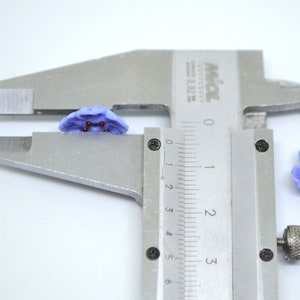 May include: Close-up of a purple flower-shaped glass bead with red accents, measured with a caliper. The bead is approximately 1 cm in diameter.