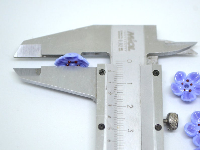May include: Close-up of a purple flower-shaped glass bead with red accents, measured with a caliper. The bead is approximately 1 cm in diameter.