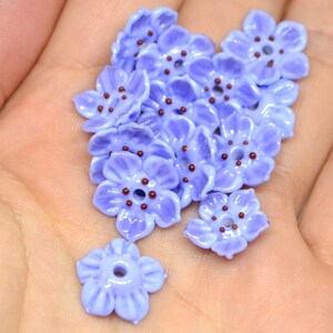 May include: A collection of 15 lavender glass flower beads with brown centers. The beads are arranged in a pile on a hand.