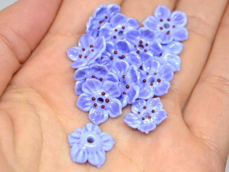 May include: A collection of 15 lavender glass flower beads with brown centers. The beads are arranged in a pile on a hand.