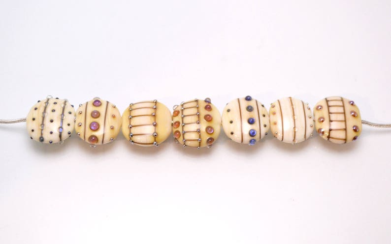 May include: A string of eight round, cream-colored beads with different patterns of brown, gold, and blue accents. The beads are strung on a silver wire.