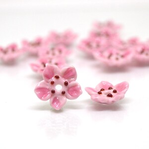 May include: Pink ceramic flower beads with a hole in the center. The beads are shaped like cherry blossoms with a red center.