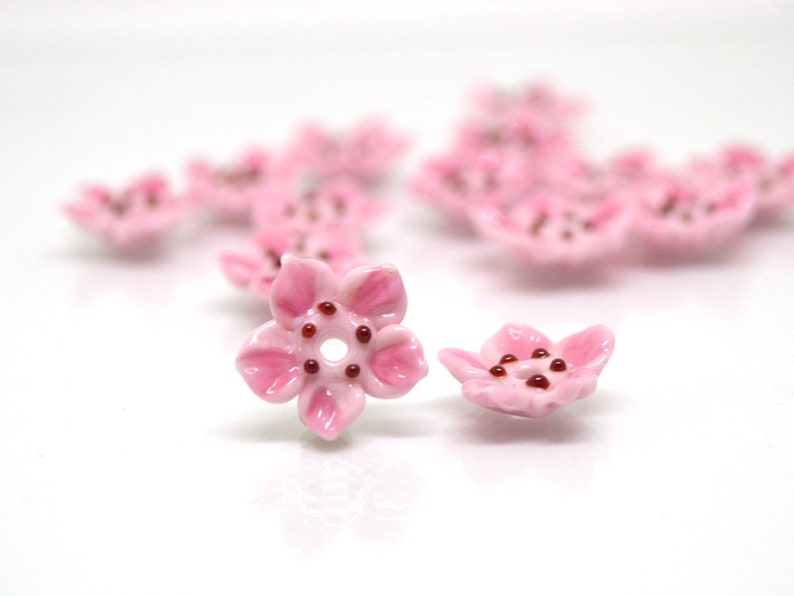May include: Pink ceramic flower beads with a hole in the center. The beads are shaped like cherry blossoms with a red center.