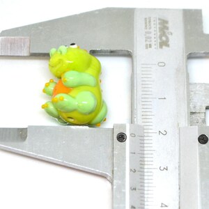 May include: A green glass bead shaped like a dinosaur with orange accents. The bead is being measured with a ruler.