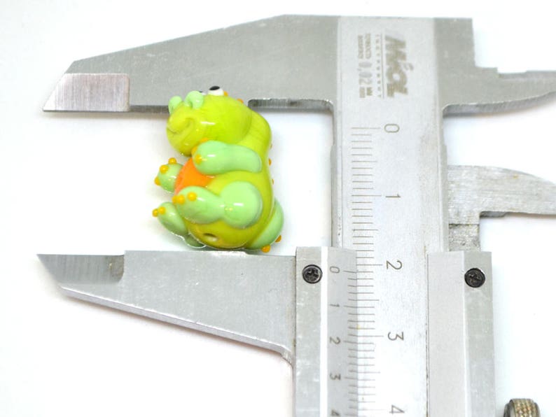 May include: A green glass bead shaped like a dinosaur with orange accents. The bead is being measured with a ruler.
