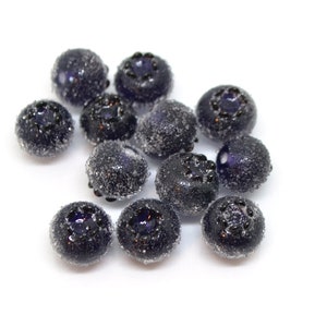 May include: A group of 12 dark blue glass blueberry beads with a frosted finish. The beads are round and have a slightly raised design on the top.