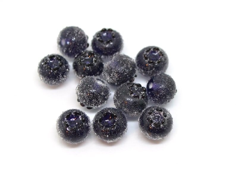 May include: A group of 12 dark blue glass blueberry beads with a frosted finish. The beads are round and have a slightly raised design on the top.