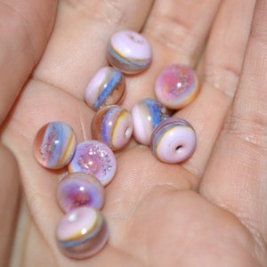 May include: A collection of iridescent glass beads in shades of pink, purple, and blue. The beads have a layered, swirled effect and are perfect for jewelry making.