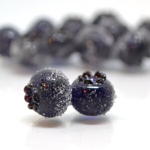 May include: Close-up of two dark blue glass beads with a frosted finish. The beads have a textured surface and are slightly out of focus. The beads are surrounded by a white background.