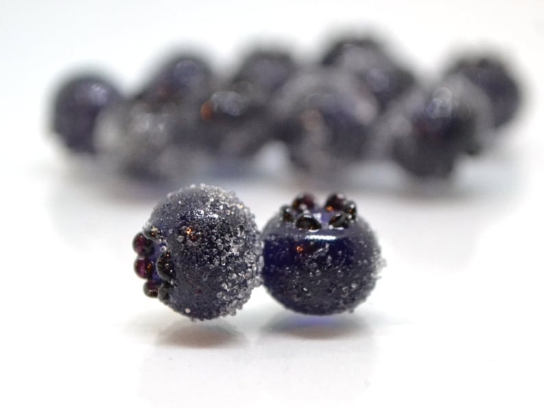 May include: Close-up of two dark blue glass beads with a frosted finish. The beads have a textured surface and are slightly out of focus. The beads are surrounded by a white background.