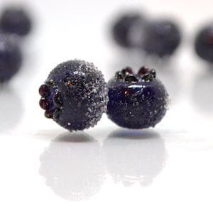 May include: Two dark blue glass blueberries with a frosted finish. The blueberries have a dark purple hue and are slightly out of focus.