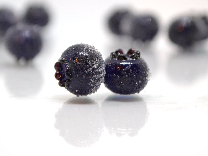 May include: Two dark blue glass blueberries with a frosted finish. The blueberries have a dark purple hue and are slightly out of focus.