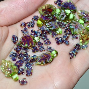 May include: A collection of glass beads in shades of green, purple, and brown. The beads are shaped like leaves and flowers.