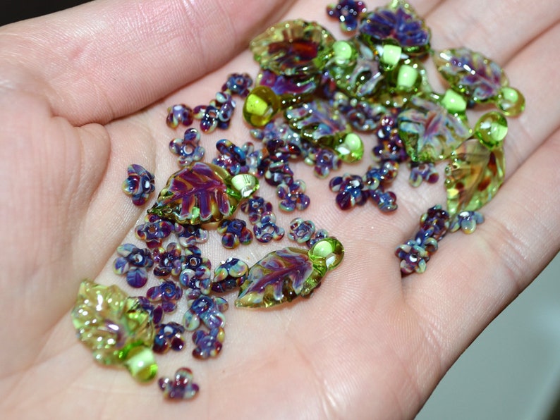 May include: A collection of glass beads in shades of green, purple, and brown. The beads are shaped like leaves and flowers.