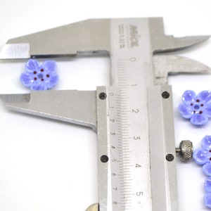 May include: Close-up of a caliper measuring the size of a purple flower-shaped bead. The bead has a five-petal design and a dark purple center.