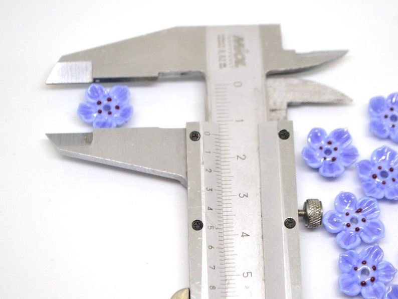 May include: Close-up of a caliper measuring the size of a purple flower-shaped bead. The bead has a five-petal design and a dark purple center.