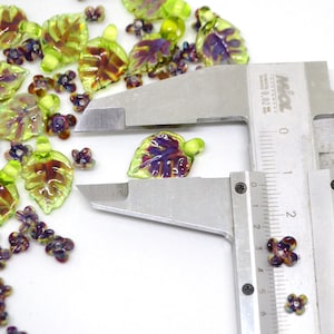 May include: Assortment of green and purple glass leaf and flower beads, measured with a ruler. The leaf beads are approximately 1.5 inches long. The flower beads are approximately 0.5 inches wide.