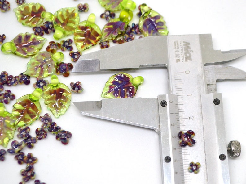 May include: Assortment of green and purple glass leaf and flower beads, measured with a ruler. The leaf beads are approximately 1.5 inches long. The flower beads are approximately 0.5 inches wide.