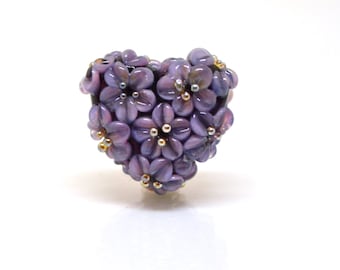 Könnte beinhalten: Eine herzförmige Glasperle mit einem Cluster aus lila Blüten mit goldenen Zentren.