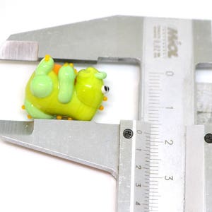 May include: A green and yellow glass figurine of a cartoon monster with orange and yellow accents. The figurine is being measured with a ruler.
