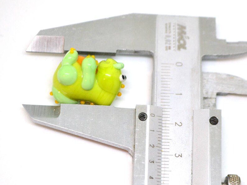 May include: A green and yellow glass figurine of a cartoon monster with orange and yellow accents. The figurine is being measured with a ruler.