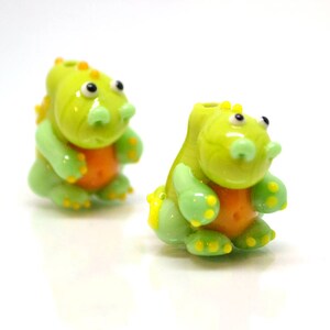 May include: Two green glass dinosaur figurines with orange accents. The dinosaurs have yellow eyes and are sitting upright.
