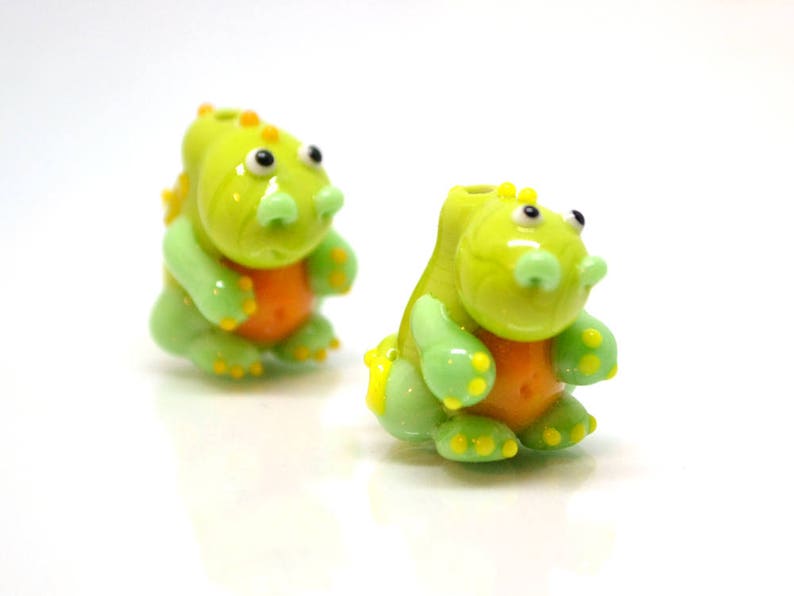May include: Two green glass dinosaur figurines with orange accents. The dinosaurs have yellow eyes and are sitting upright.