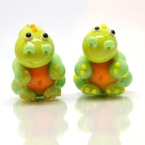 May include: Two small green glass dinosaur figurines with orange bellies and yellow accents. The dinosaurs are sitting on a white surface.