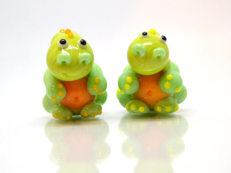 May include: Two small green glass dinosaur figurines with orange bellies and yellow accents. The dinosaurs are sitting on a white surface.