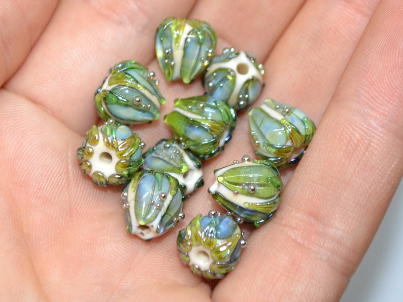 K&ouml;nnte beinhalten: Eine Sammlung von neun gr&uuml;nen und wei&szlig;en Glasperlen mit aufwendigen Designs. Die Perlen haben eine perlmuttartige Oberfl&auml;che und sind mit silbernen Punkten verziert.