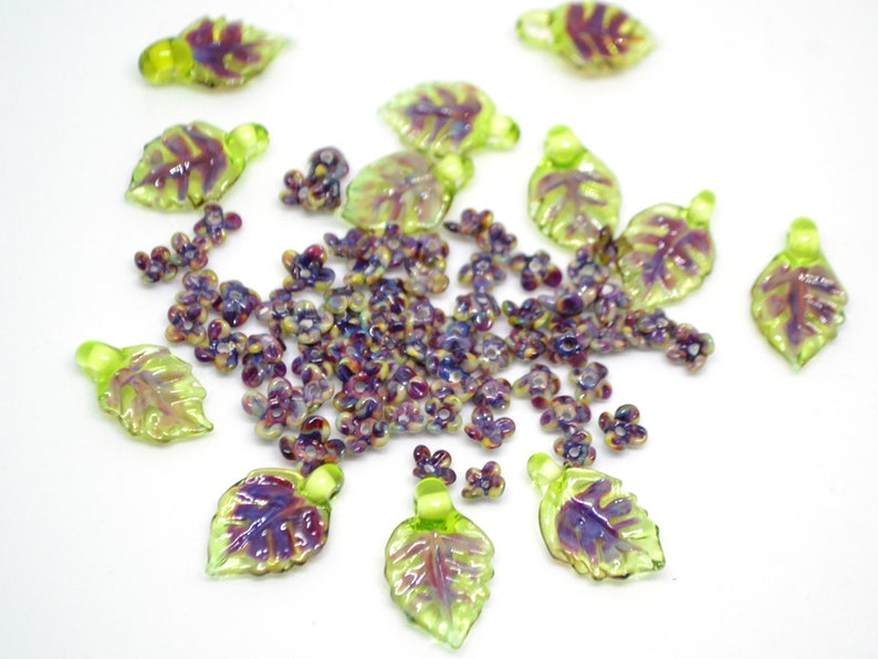 May include: Assortment of green and purple glass leaf charms with iridescent accents. The charms are scattered on a white surface.
