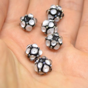 May include: Six black and white glass beads with a textured surface. The beads are round and have a small hole in the centre.