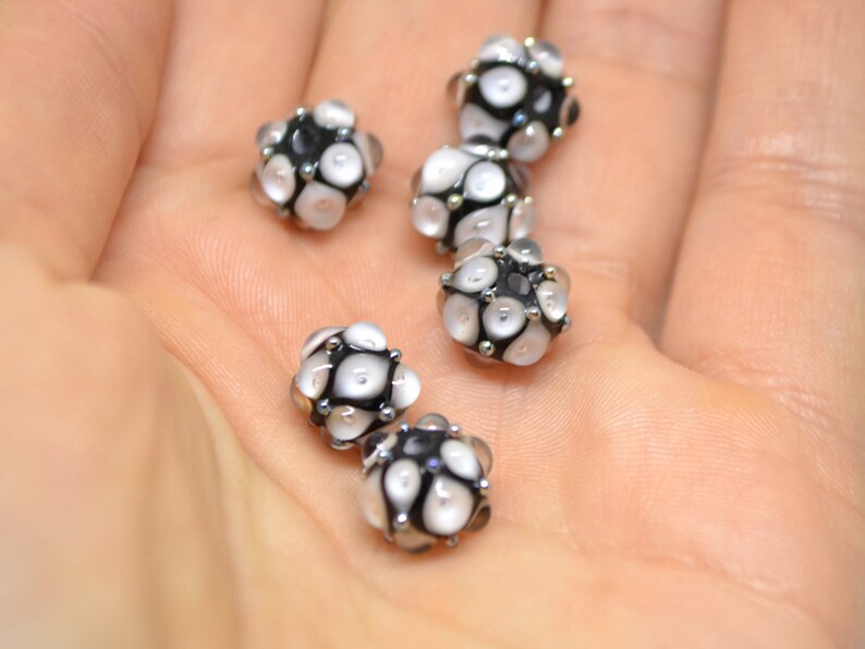 May include: Six black and white glass beads with a textured surface. The beads are round and have a small hole in the centre.