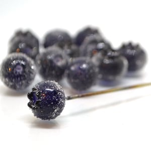 May include: A cluster of small, dark blue glass beads with a frosted finish, resembling blueberries. The beads are arranged on a white surface with a thin, gold-colored wire running through them.