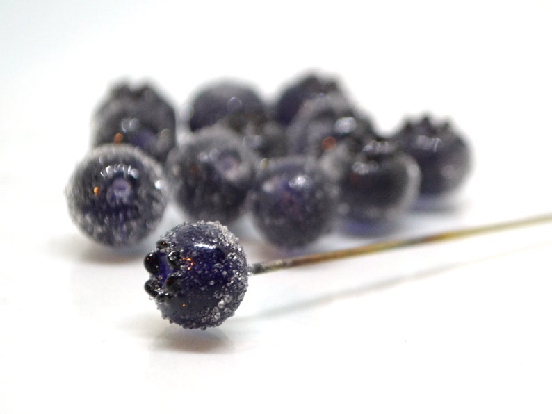 May include: A cluster of small, dark blue glass beads with a frosted finish, resembling blueberries. The beads are arranged on a white surface with a thin, gold-colored wire running through them.
