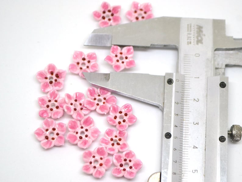 May include: Pink ceramic flower beads with a red center, measured with a ruler. The beads are approximately 1 cm in diameter.