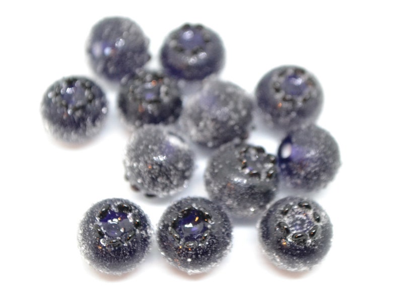 May include: A close-up of a group of dark blue glass beads with a frosted finish. The beads are round and have a slightly irregular shape. They are scattered on a white surface.