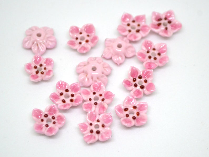 May include: Pink ceramic flower beads with a light pink glaze and dark pink centers. The beads are shaped like five-petal flowers.